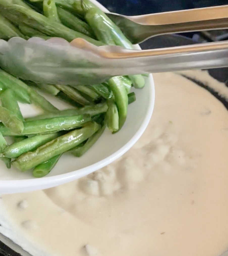 Adding cooked green beans with silver tongs to sour cream and soup mixture.