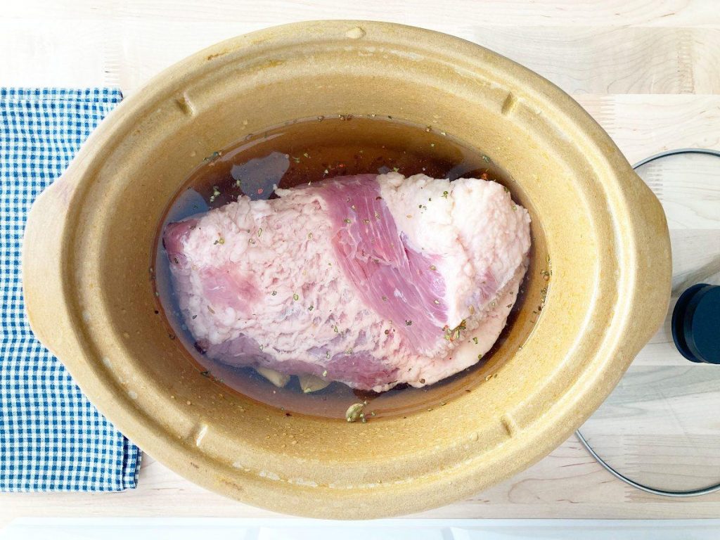 Water filled above uncooked cornbeef in crockpot, white and blue checkered towel and crockpot cover.
