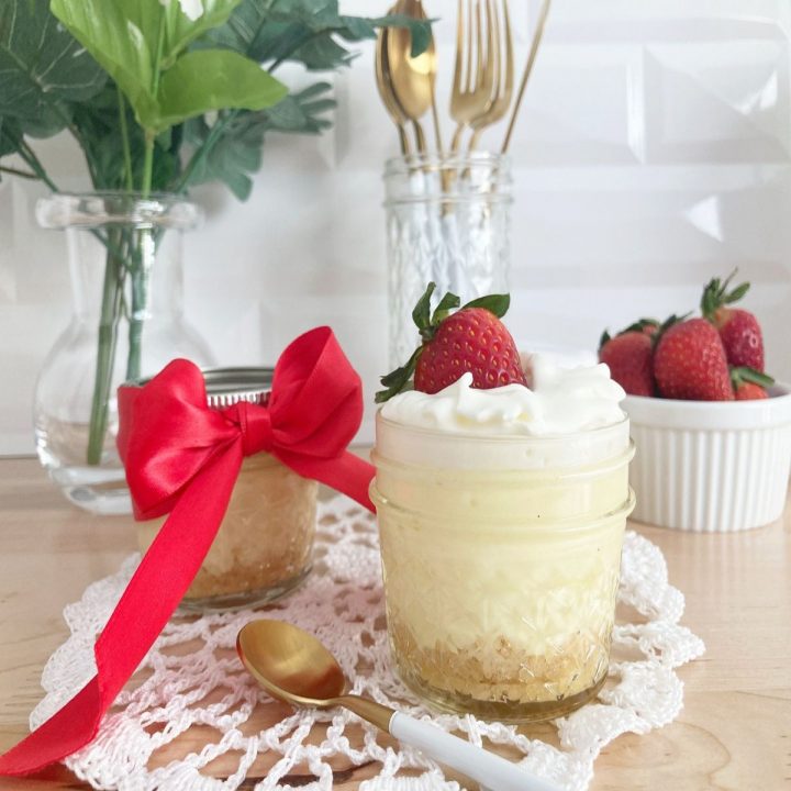 Finished new york cheesecake jar topped with whipped cream and strawberry. Next to red ribbon decorated closed cheesecake jar and bowl of strawberries and utensils in background.