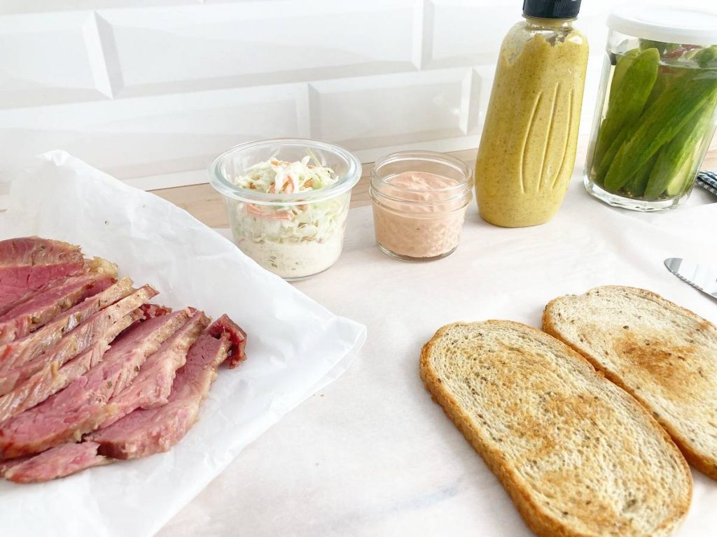 Sliced cornbeef on parchment, glass jar with coleslaw, glass jar with russian dressing, bottle of spicy deli mustard, glass jar with fresh homemade pickles, and two slices of dry rye bread.