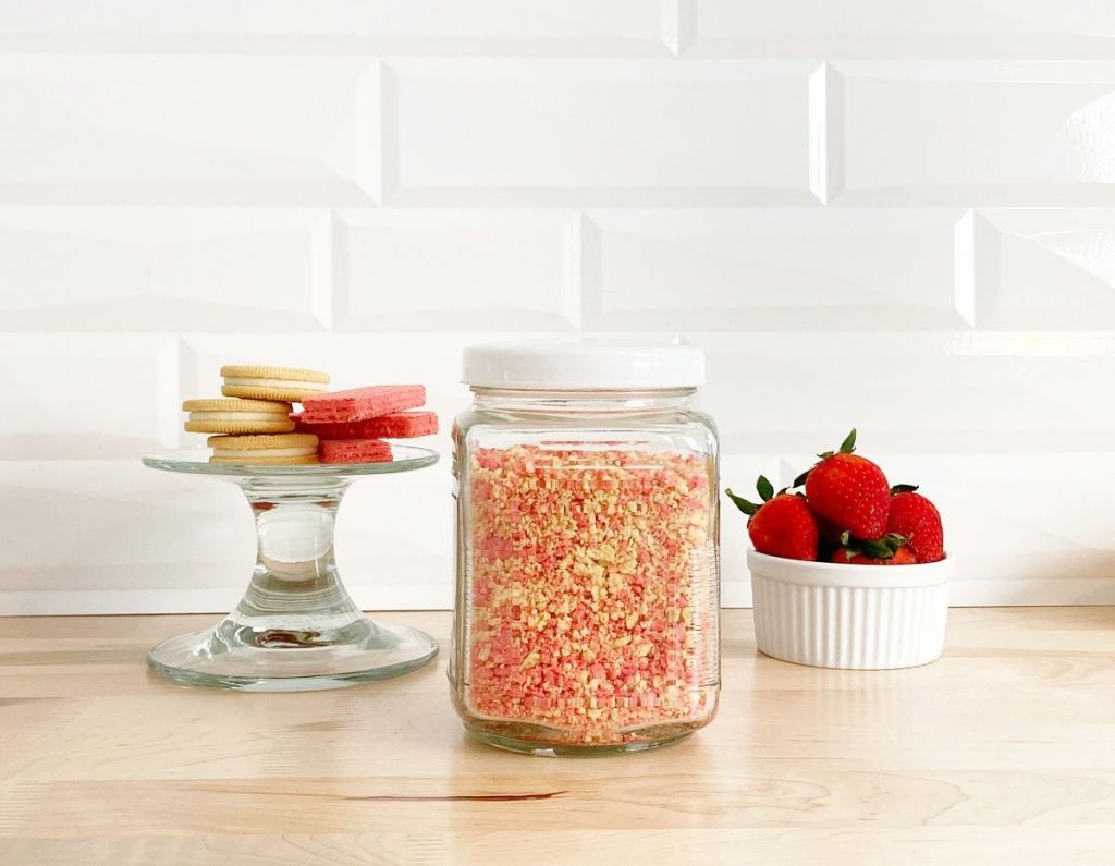 Strawberry shortcake crumble stored in jar. Next to bowl of fresh strawberries, golden oreos and wafers.