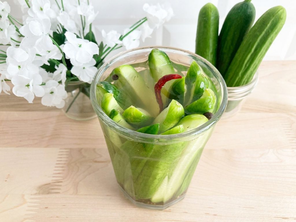 Finished pickled cucumber before putting into refrigerator, with no lid next to white flowers and 3 whole mini cucumbers.