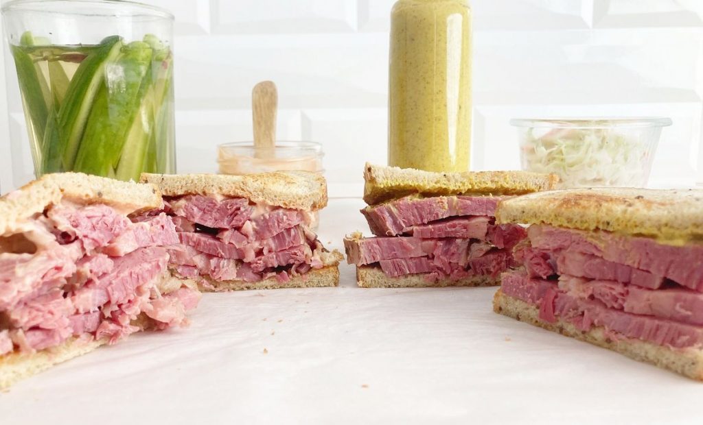 Both cornbeef sandwiches sliced in half, glass jar with coleslaw, glass jar with russian dressing, bottle of spicy deli mustard, glass jar with fresh homemade pickles.