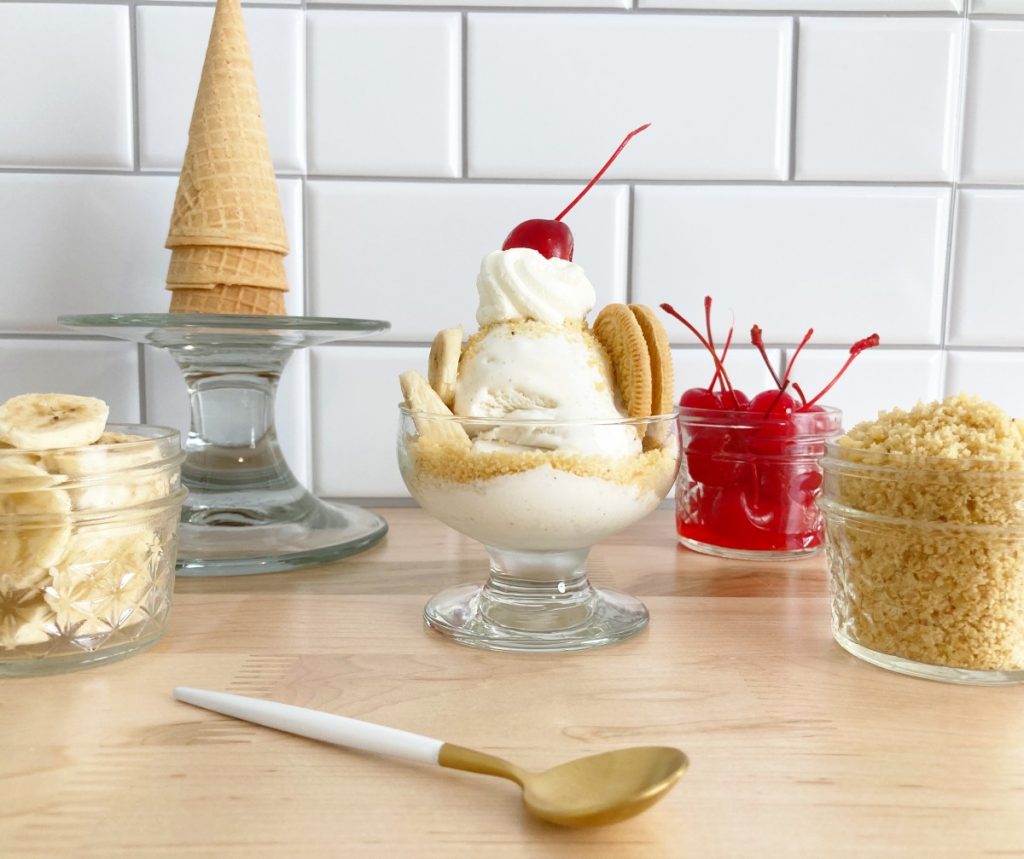 Banana pudding crunch sundae. Next to small mason jars filled with fresh sliced bananas, cherries, extra crumbles, and waffle cones.