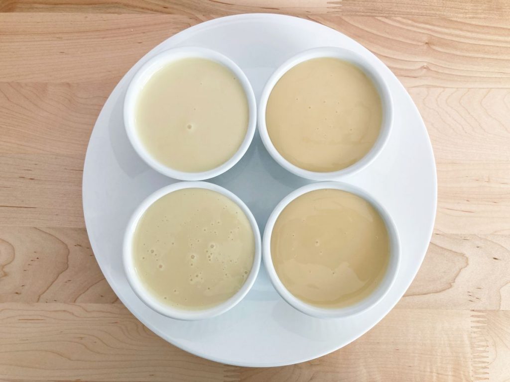 Four white ramekins filled with condensed milk on white plate. Wood background.