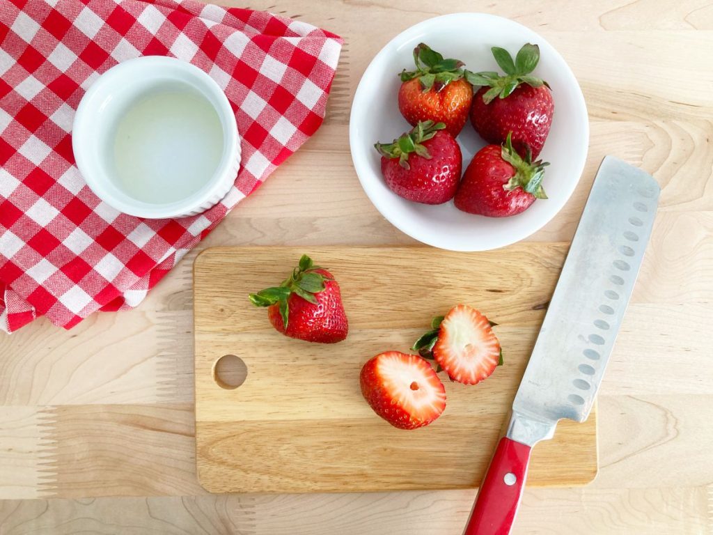Cutting tops of strawberries on wood cutting board with red knife.