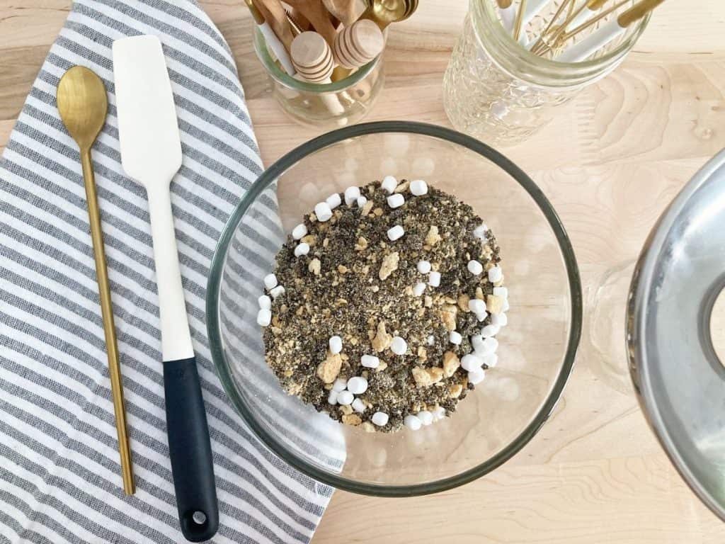 Finished bowl of s'more crunch topping next to mason jar and funnel.Next to white spatula, gold spoon, and grey white striped towel.