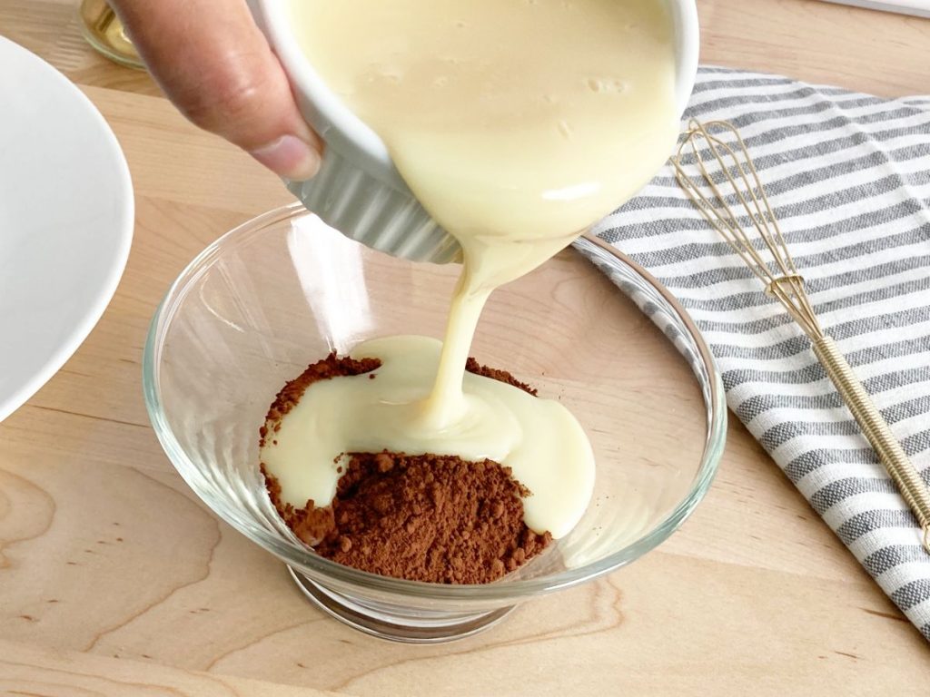Pouring condensed milk into glass bowl with cocoa powder. Next to gold whisk and grey white striped towel.