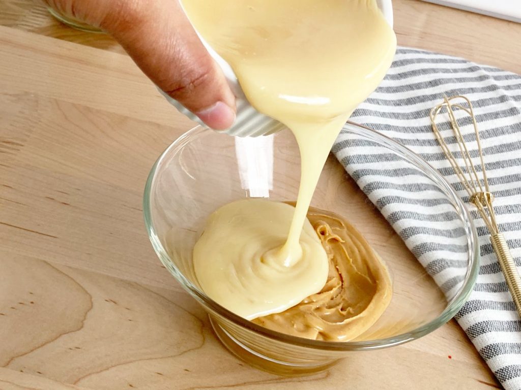 Pouring condensed milk into glass bowl with peanut butter. Next to gold whisk and grey white striped towel.