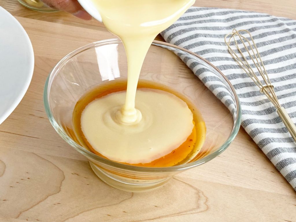 Pouring condensed milk into glass bowl with vanilla extract. Next to gold whisk and grey white striped towel.
