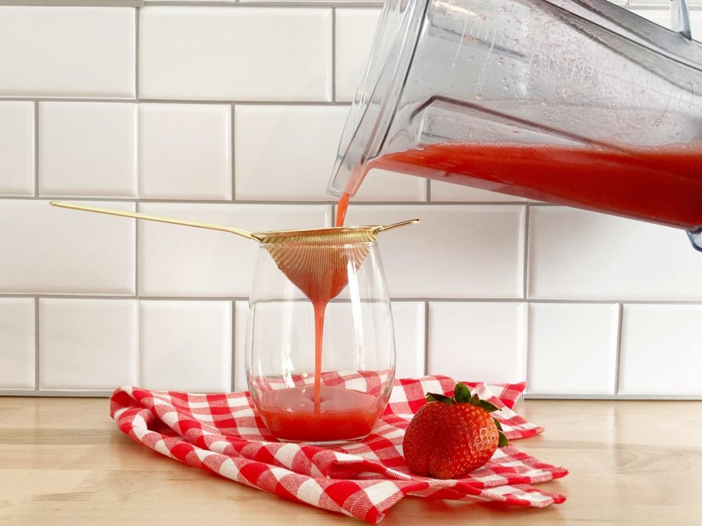 Pouring finished strawberry juice into a gold strainer on top serving glass cup.
