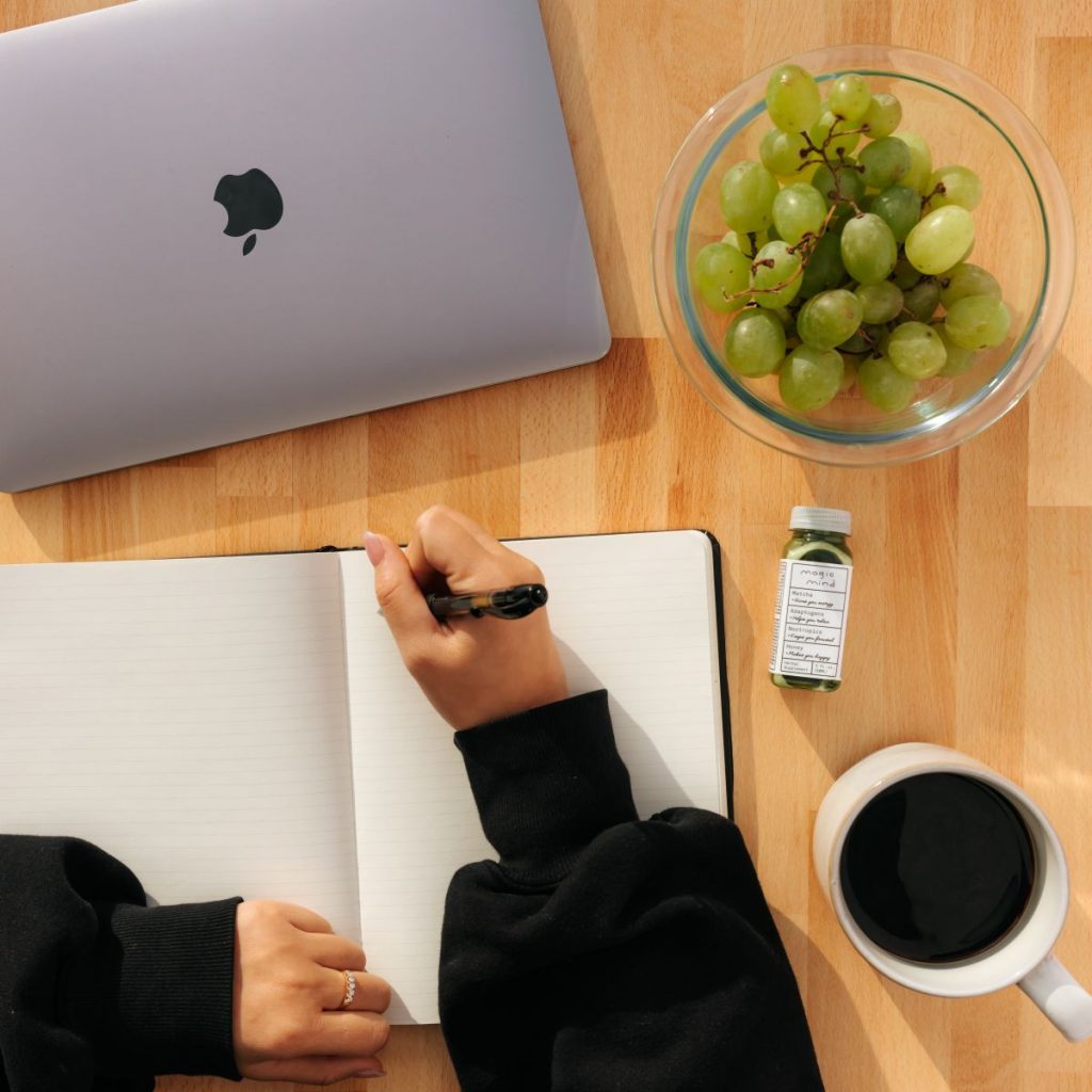 Hands writing in a notebook next to a cup of coffee, MacBook, and grapes. Great place to start broke grocery list.