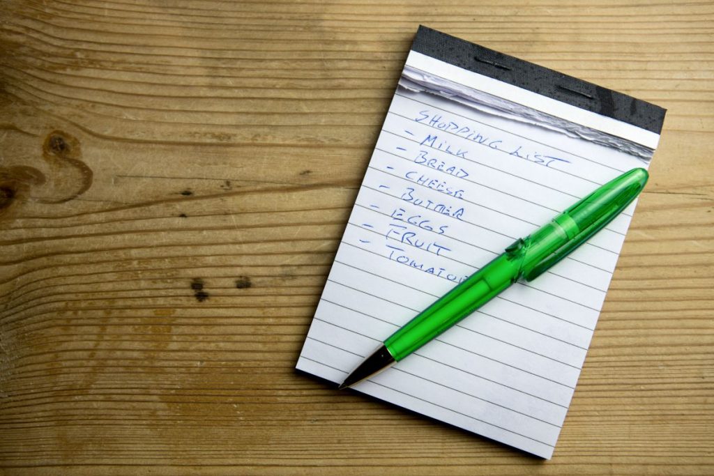 Notepad with a grocery shopping list and green pen on wood background.