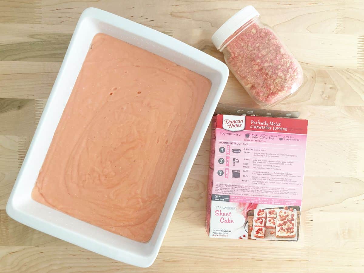 Unbaked strawberry boxed cake mix batter in a white baking dish. Next to empty box to strawberry cake mix and jar of strawberry crunch crumbles.