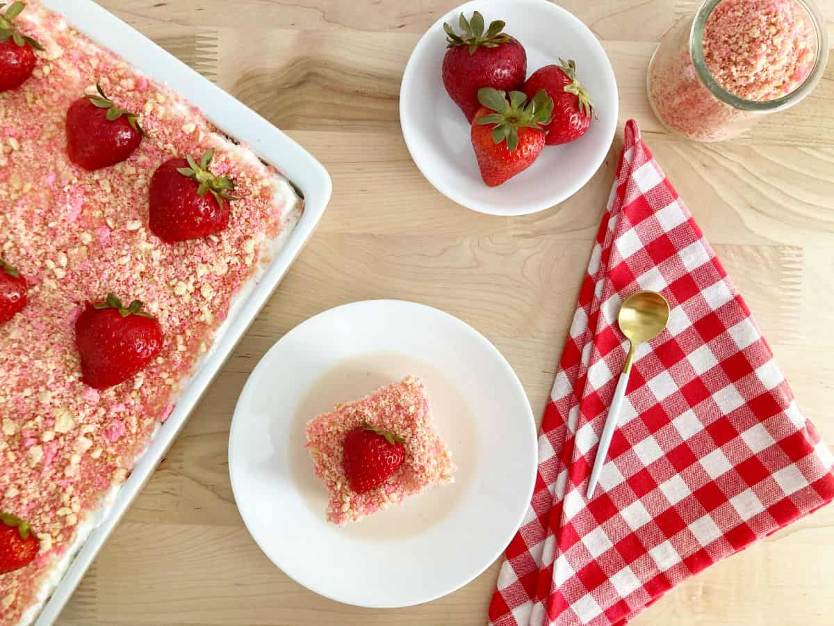 Strawberry crunch tres leche cake in white dish topped with sliced fresh strawberries. A slice of cake, bowl of strawberries, gold spoon and red white checkered napkin.