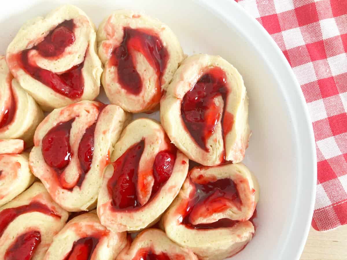Uncooked strawberry rolls in white pie pan next to white red checkered kitchen towel