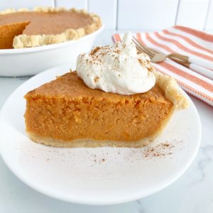 Close up slice of condensed milk sweet potato pie topped with whipped cream and sprinkled with spice on white plate. Behind it is whole cut pie, gold fork and knife and orange white kitchen towel.