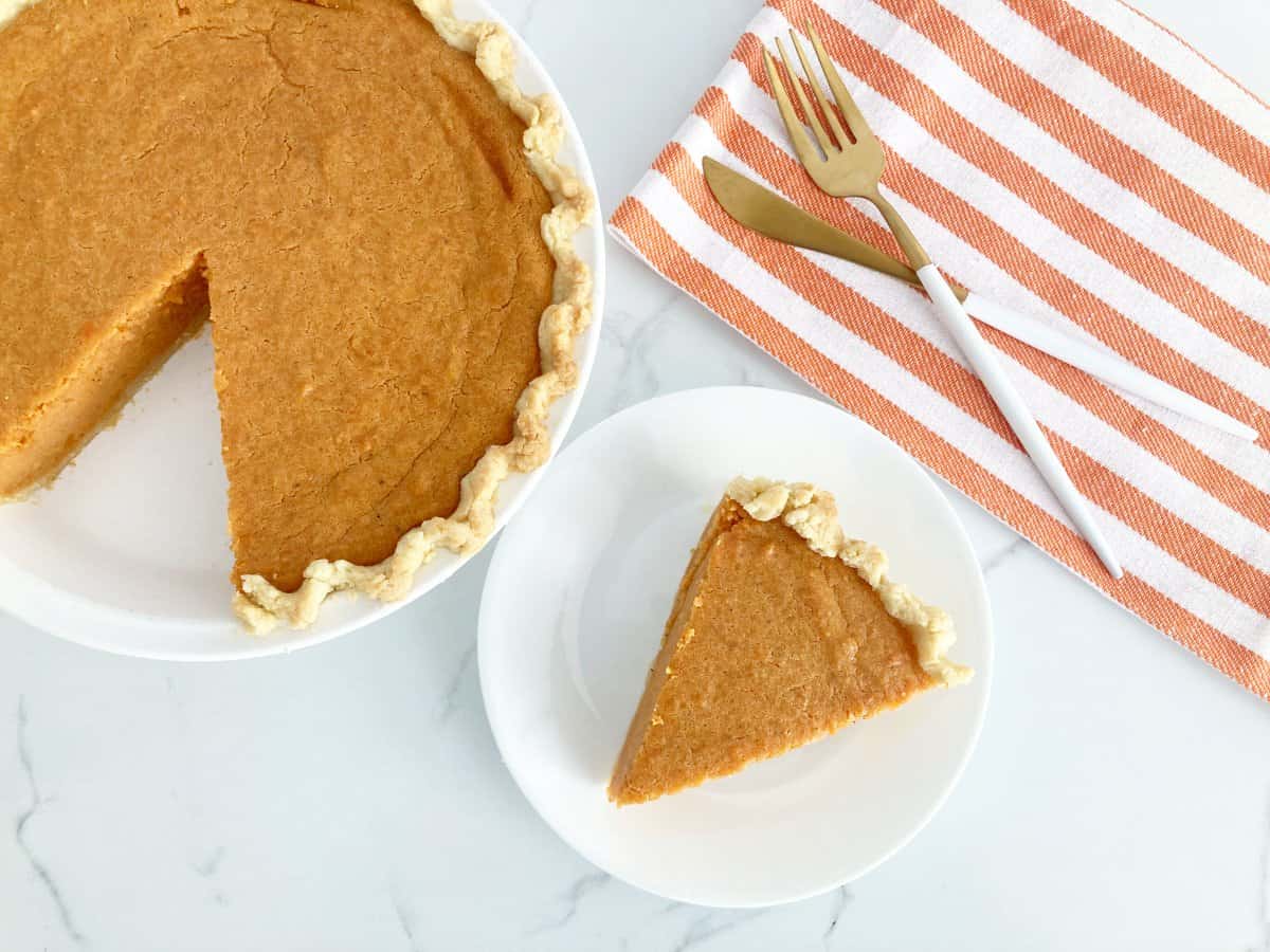 Whole cut brown butter sweet potato pie next to a plate with a slice of pie. Next to the pie is gold knife and fork on top of orange and white kitchen towel.