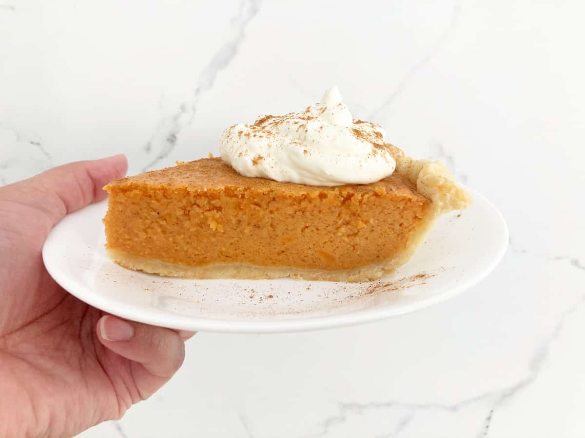 Hand holding slice of brown butter sweet potato pie topped with whipped cream and sprinkled with mixed spice on white plate.