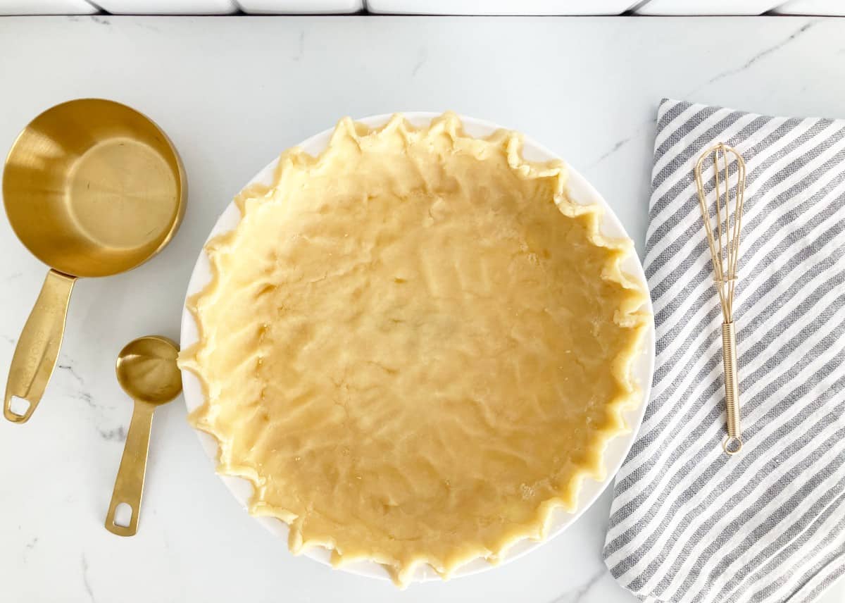 Unbaked pie crust next to small gold whisk, gold measuring cup, gold measuring spoon, and grey white kitchen towel on marble counter top.