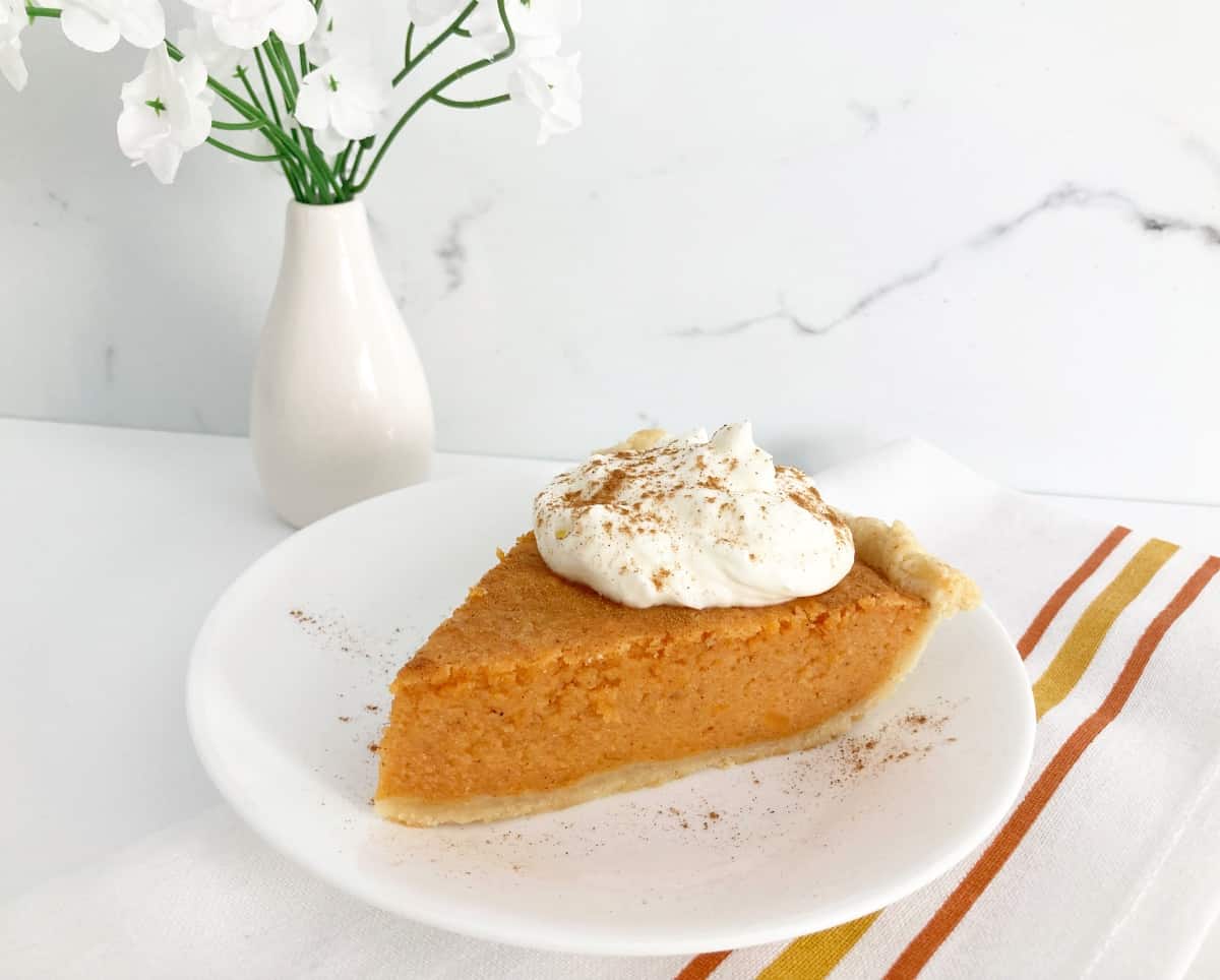 Slice on pie on white plate topped with whipped cream and mixed spice. Next to orange and white napkin and white vase with white flowers.