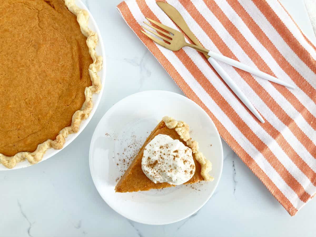 Whole cut brown butter sweet potato pie next to a plate with a slice of pie topped with whipped cream and sprinkled with mixed spice. Next to the pie is gold knife and fork on top of orange and white kitchen towel.