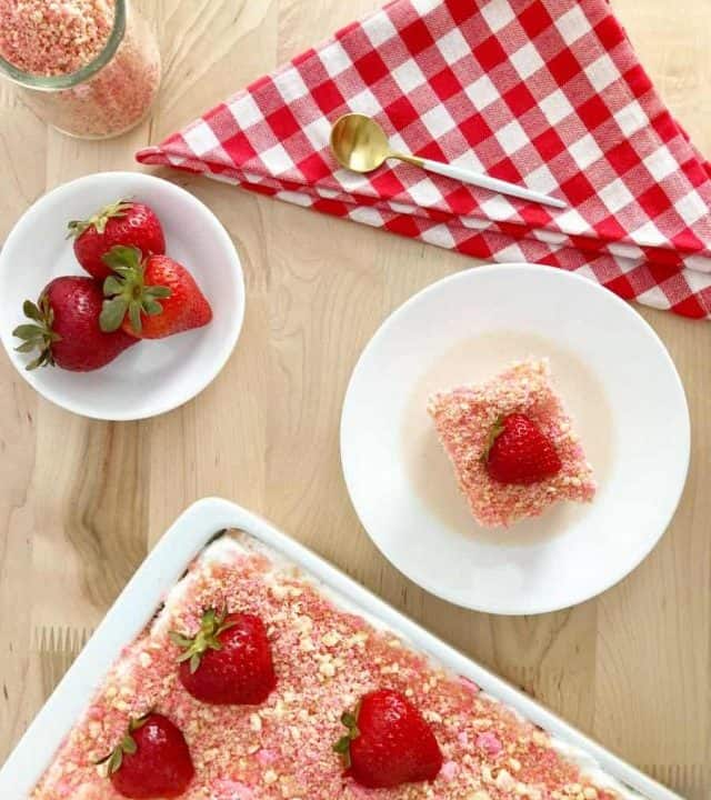 Strawberry crunch tres leche cake in white dish topped with sliced fresh strawberries. A slice of cake, bowl of strawberries, a gold spoon, and red white checkered napkin.
