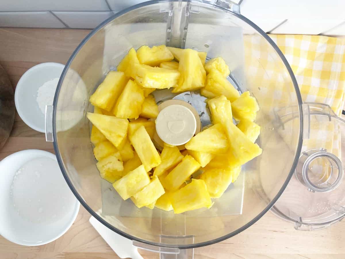 Top down view of fresh pineapple tidbits in food processor jar with ingredients and lid next to it.