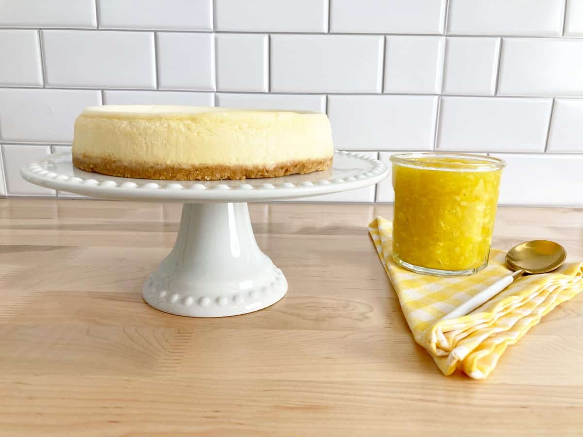 Side of view of a plain cheesecake on a white cake stand next to glass jar with the topping and a gold spoon.