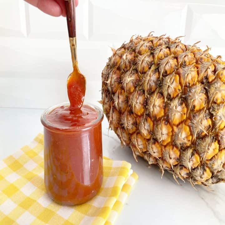 Small clear jar of pineapple bbq sauce with a hand holding small gold spoon showing the texture of the sauce. Next to it is a ripe fresh pineapple.