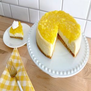 A slice of cheesecake with the pineapple topping on a white plate. Next to the whole cheesecake with the topping.