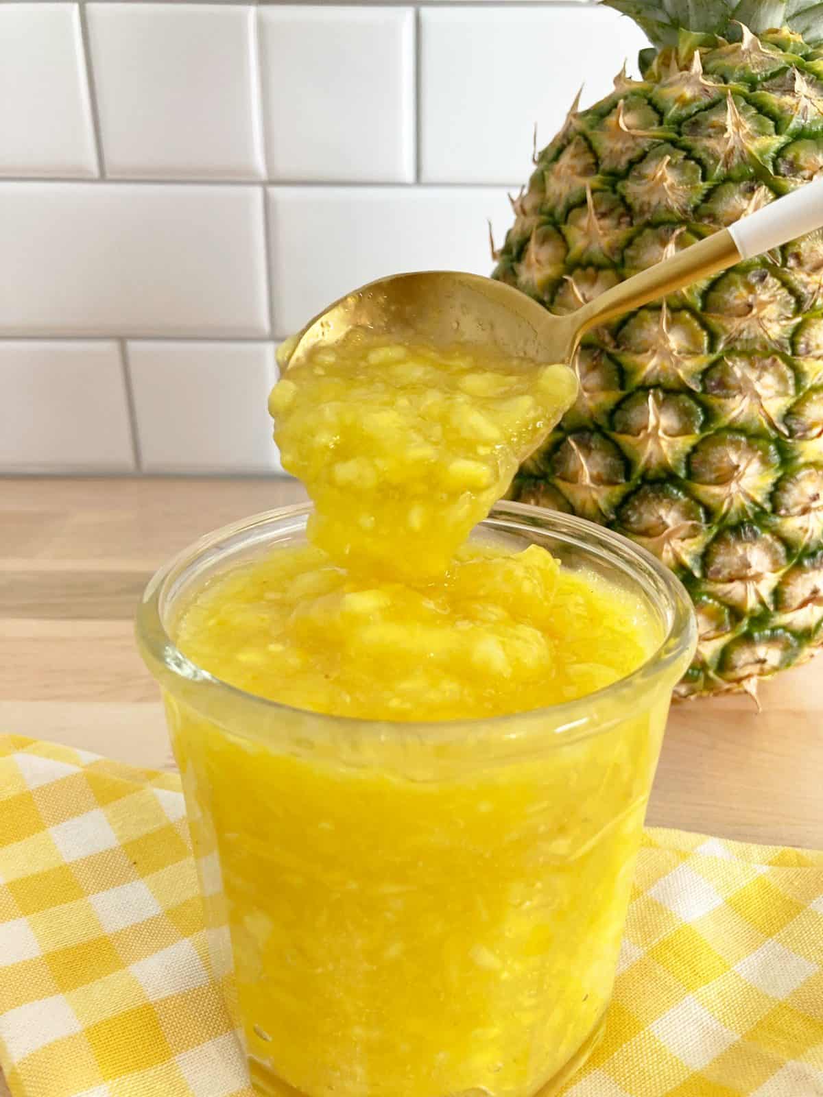 Glass jar filled with pineapple topping and a gold spoon showing the texture. Behind is a whole fresh pineapple.