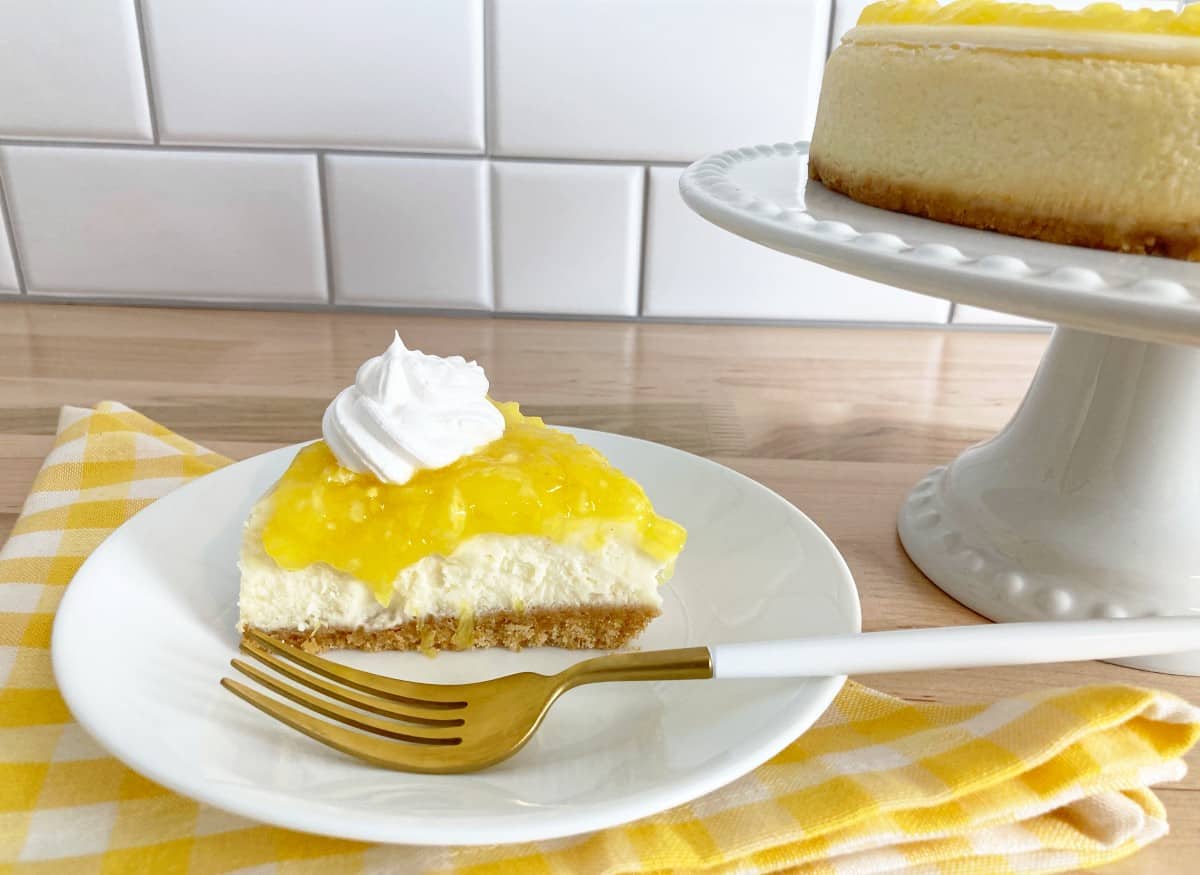 Slice of pineapple cheesecake with whipped cream on a white plate and gold fork. Next to a whole pineapple cheesecake on a white cake stand.
