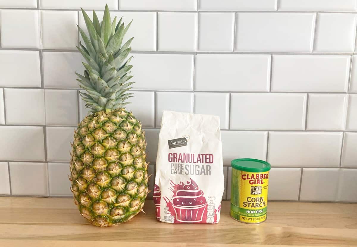Whole fresh unpeeled pineapple next to bag of white sugar and canister of corn starch.