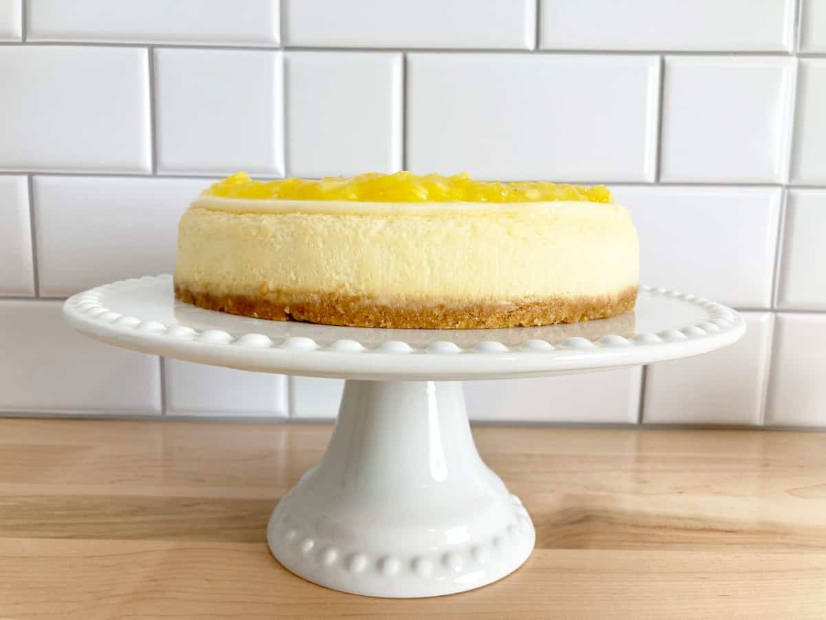 Plain cheesecake with pineapple topping on a white cake stand.