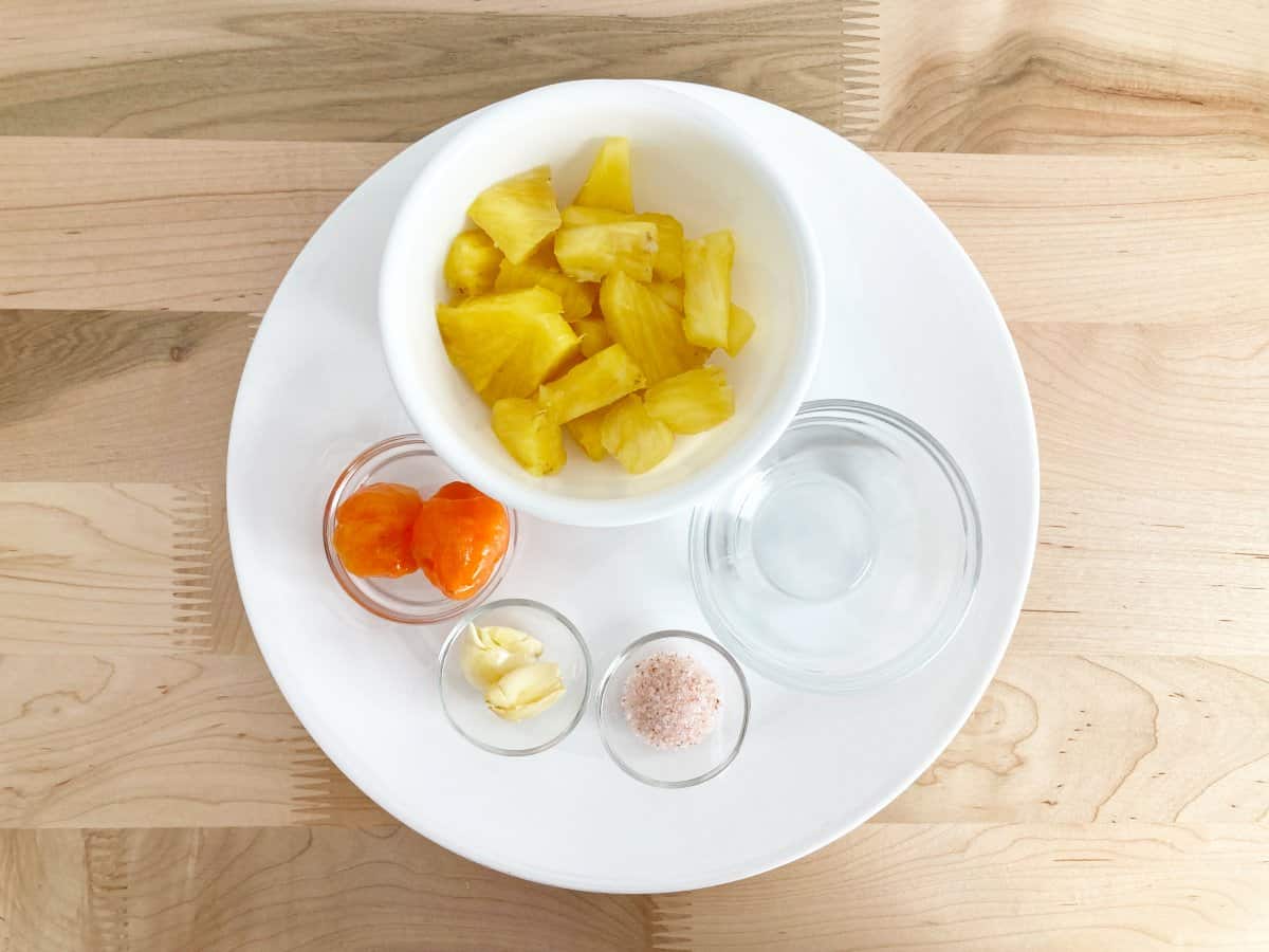 All ingredients on a white plate. Fresh pineapples, orange habanero peppers, garlic, salt, and vinegar.
