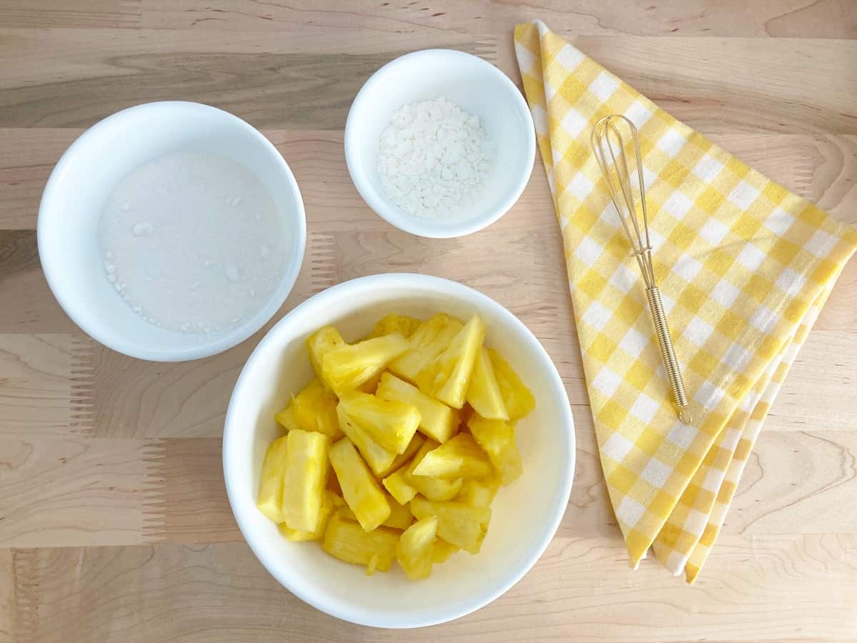 Topping ingredients including chopped fresh pineapple, white sugar, and cornstarch prepped in white bowls. Next to yellow and white checkered towel and gold whisk.