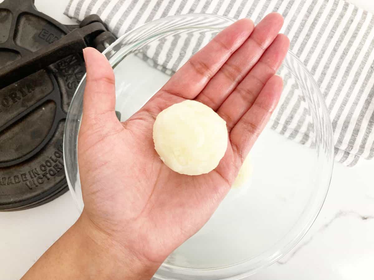 Hand holding a cassava dough ball. Next to a glass bowl, cast iron tortilla press and white and grey towel.