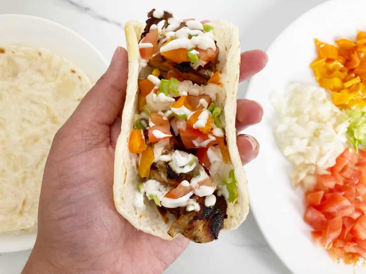 Hand holding chicken taco dressed with tomato, onions, pepper, and sour cream on a cassava tortilla.