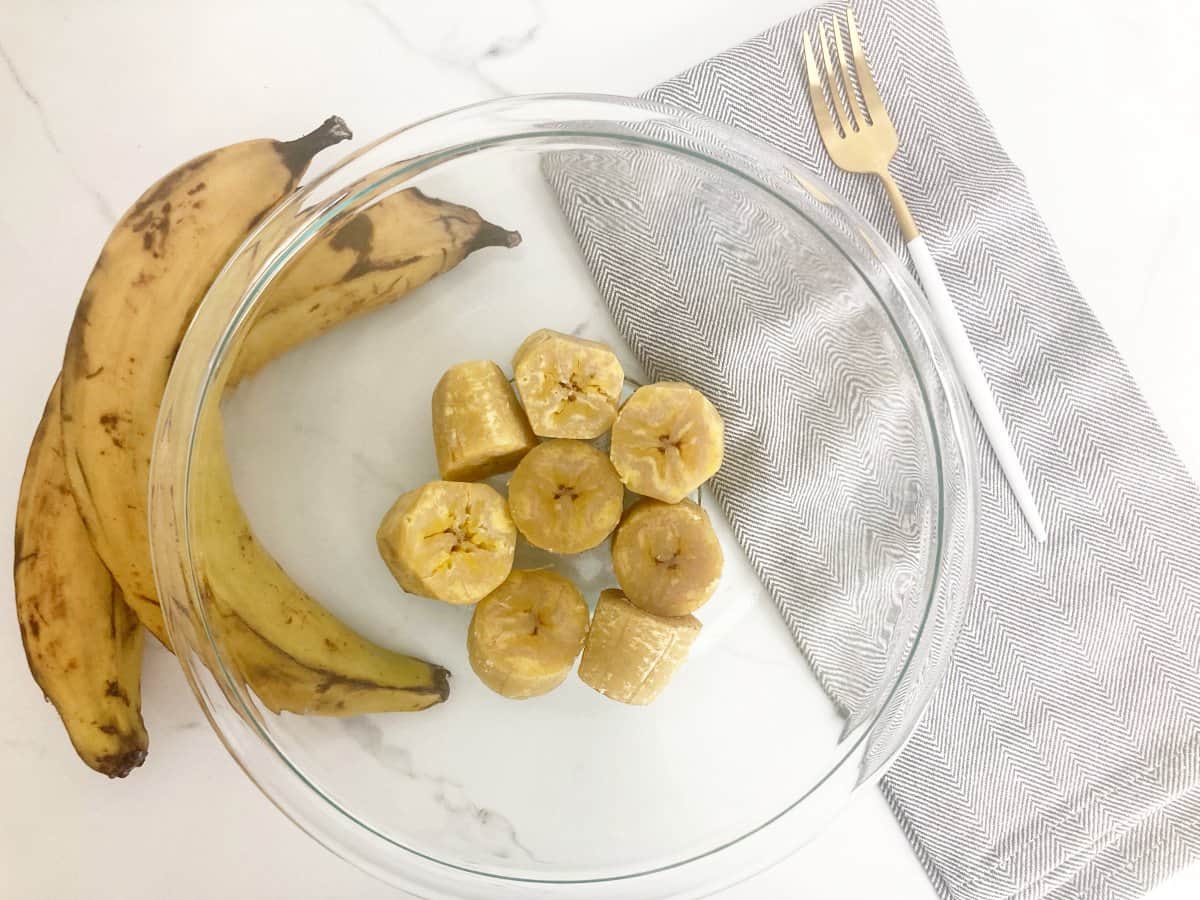 Boiled plantain chunks in glass bowl. Next to a fresh plantain, gold fork, and grey towel.