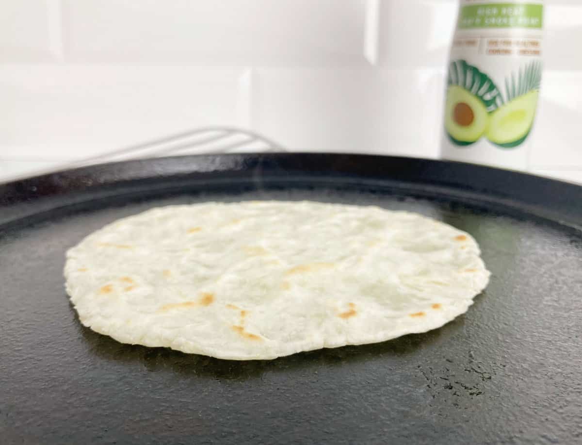 Cooking cassava tortilla on cast iron griddle. Just flipped, next to a silver metal spatula and cooking spray.