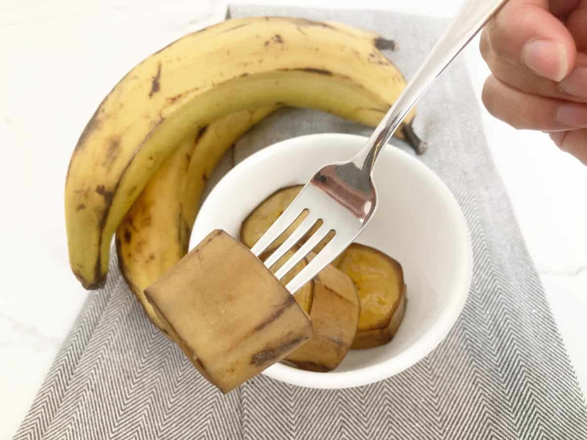 Hand holding a fork with a piece of cooked plantain with the skin on. Next to it is a white bowl of cooked plantain, two fresh plantains and a grey towel.