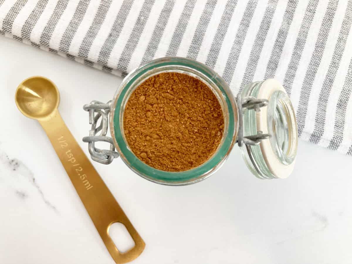 Mixed spice in a jar next to gold measuring spoon and grey and white towel.