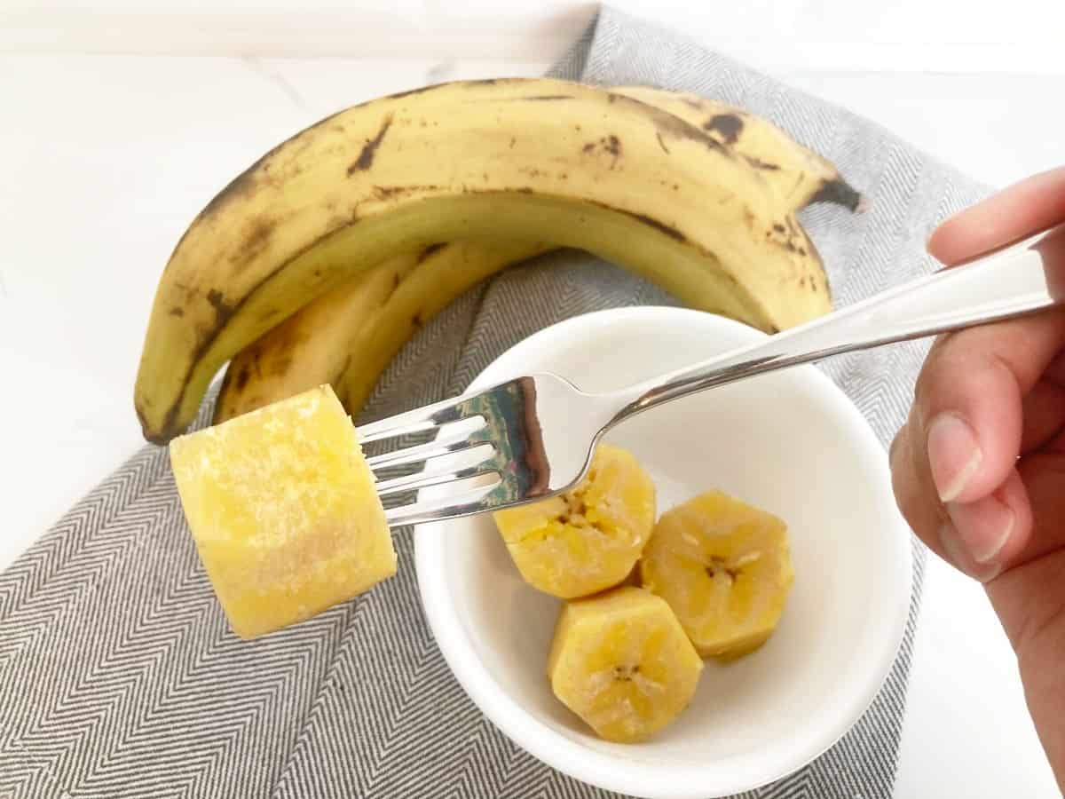 Hand holding a fork with a piece of cooked plantain. Next to it is a white bowl of cooked plantain, two fresh plantains and a grey towel.