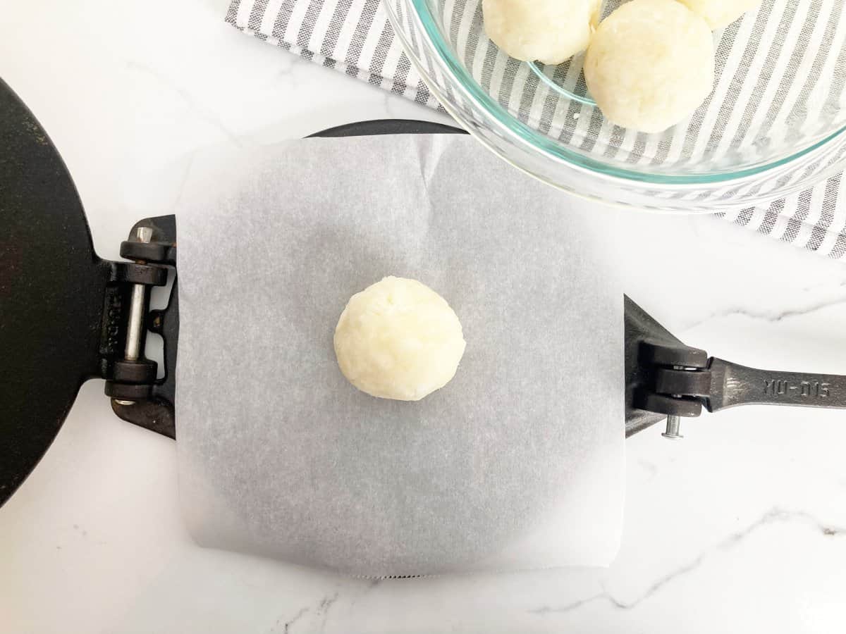 Cassava dough ball on tortilla press with parchment paper. Next to a glass bowl with portioned cassava balls on a grey and white towel.