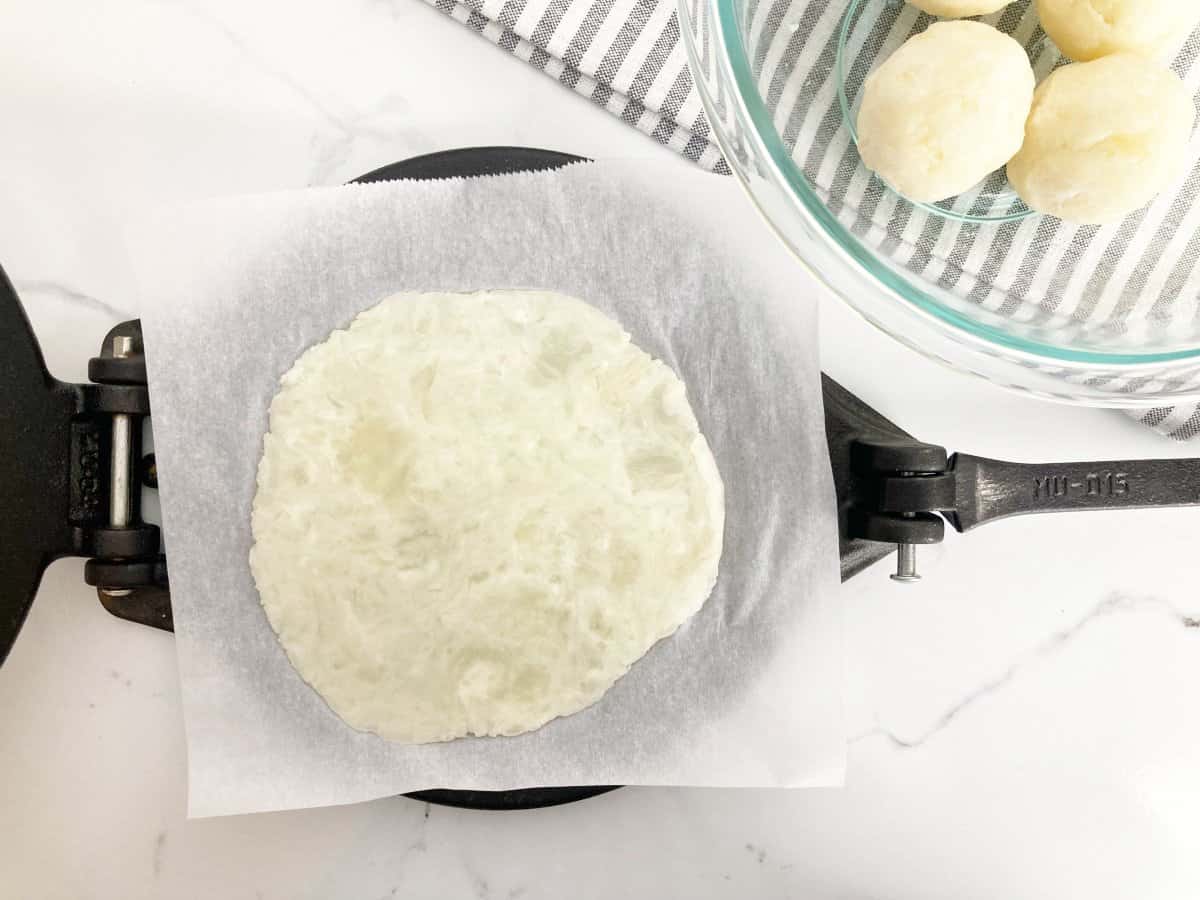 Flattened cassava tortilla dough on the tortilla press with parchment paper. Next to a bowl of cassava ball dough.