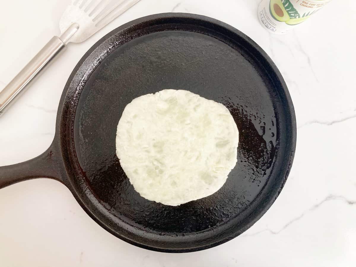 Placed flattened cassava tortilla on the hot griddle, ready to be flipped. Next to a silver metal spatula and cooking spray.