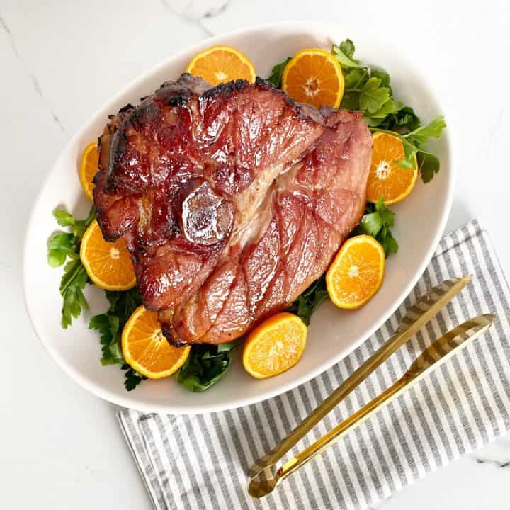 Honey baked ham on a white platter dressed with herbs and orange slices. Next to gold serving tongs on grey a white kitchen towel.