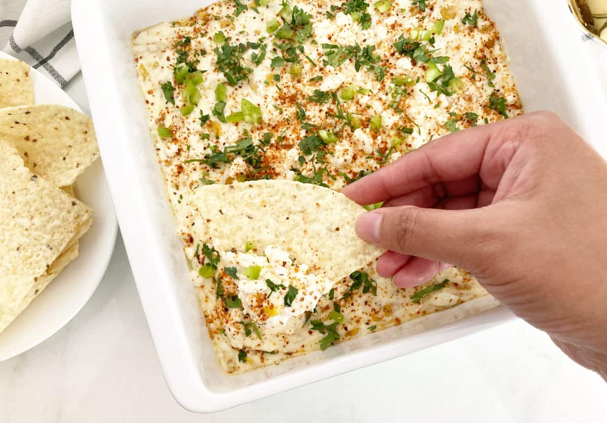 Hand holding a tortilla chip with the corn dip. Next to a baking dish of the dip and tortilla chips.