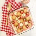 Strawberry banana pudding with shortcake crumbles. Next to a gold serving spoon, red and white checkered kitchen towel, and extra strawberry crunch topping.